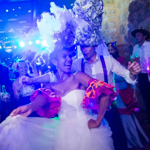A couple dancing at a lively event in Mallorca, dressed in colorful attire with feathered headpieces and surrounded by vibrant lighting