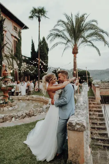 A newlywed couple embracing in a romantic finca garden setting at finca Son Tugores after their destination wedding in Mallorca, with string lights, palm trees, and a scenic view.