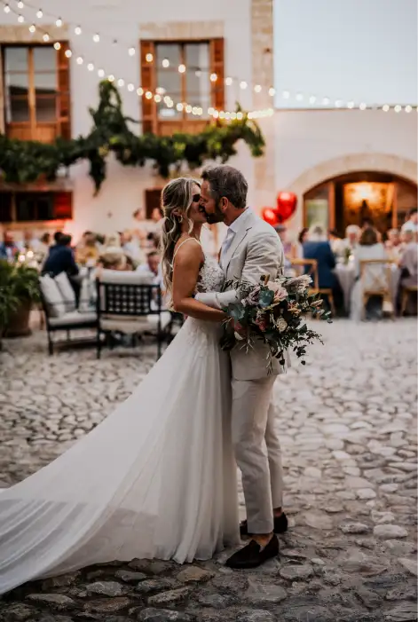 The newlyweds share a romantic kiss after their elopement in Mallorca, surrounded by string lights and a rustic courtyard setting at Las Barracas